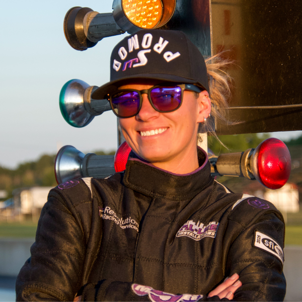 A smiling woman in a black racing suit and black cap with purple lettering stands in front of traffic lights at a racetrack. The cap reads 'PROMOD' and the racing jacket has visible sponsor patches.