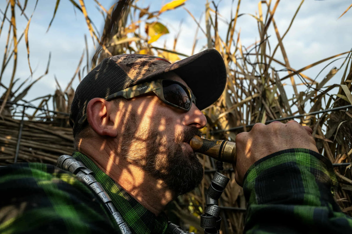 Man in camouflage gear blowing a hunting call in a grassy field